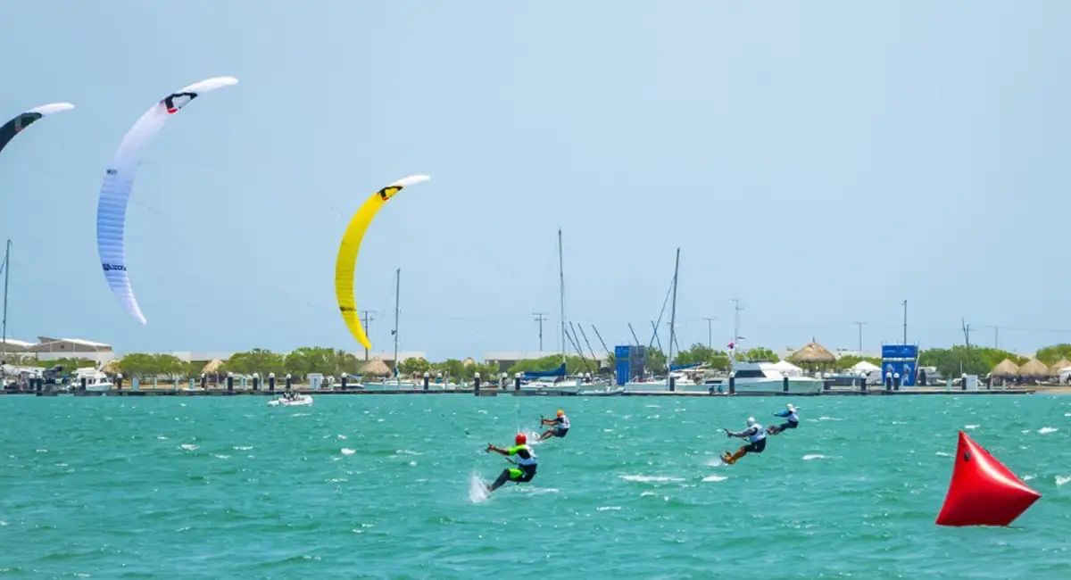 Corredores de wing foil compitiendo cerca de una boya roja en Puerto Velero, con velas blanca y amarilla al viento